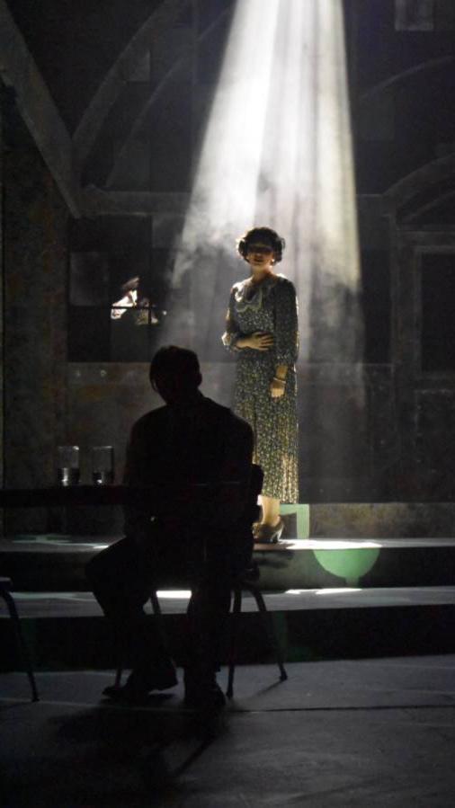Image of lady standing in dark room with a ray of sunlight shining down on her while the sillouett of a man is sitting in front of her facing away, 低着头